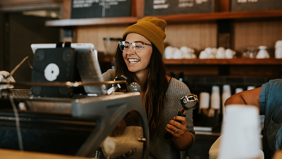 A young woman wearing glasses and a cap making coffee in a coffee shop