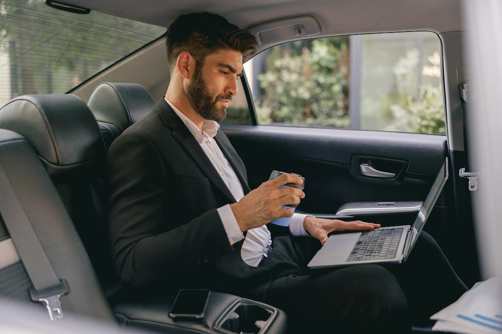 Clean-cut & professional man in suit in the back of a car, with his laptop propped up on his lap.