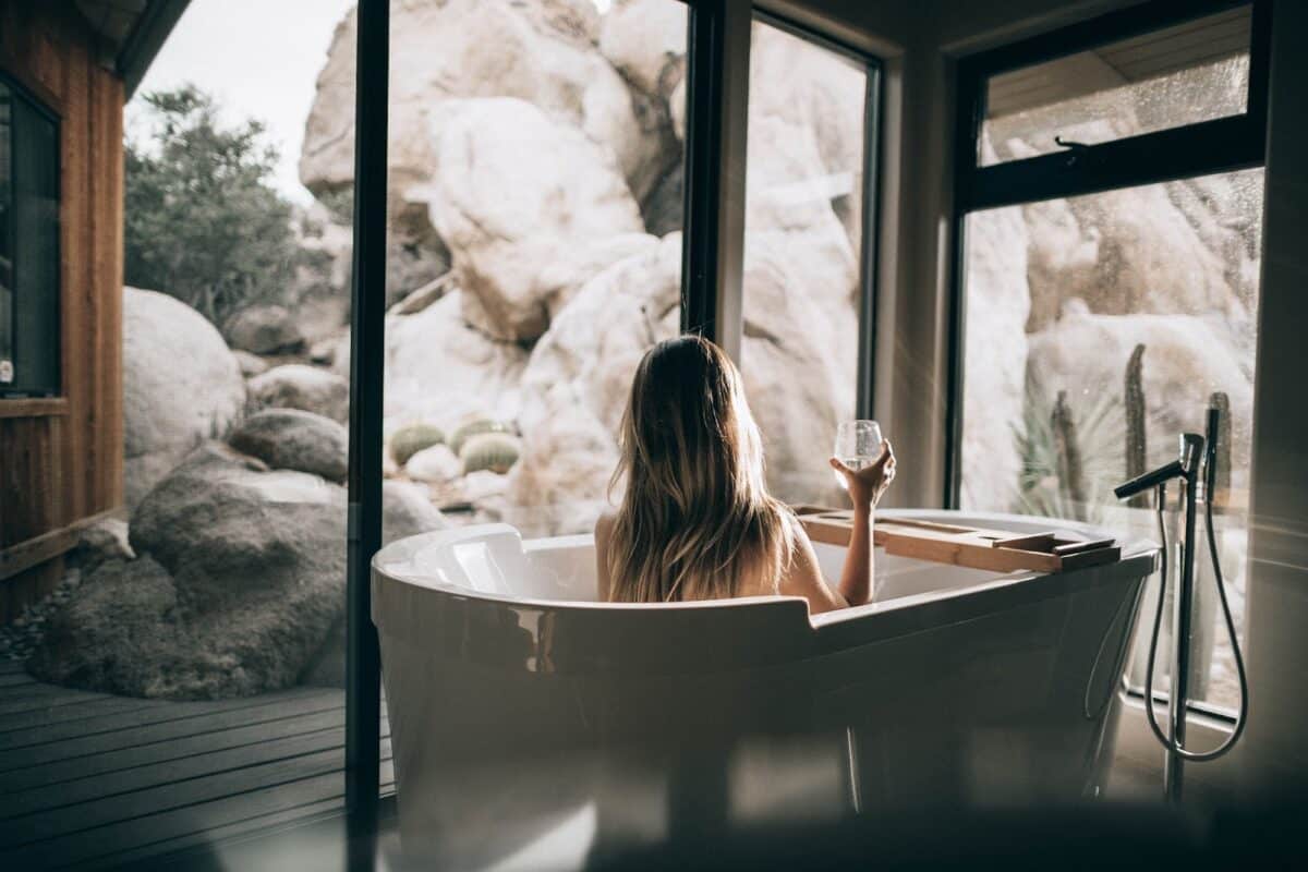 a woman faces out to a big window showing nature, while in a big soaking tub drinking a glass of water, which reflects the interior design trends, including prioritizing wellness.