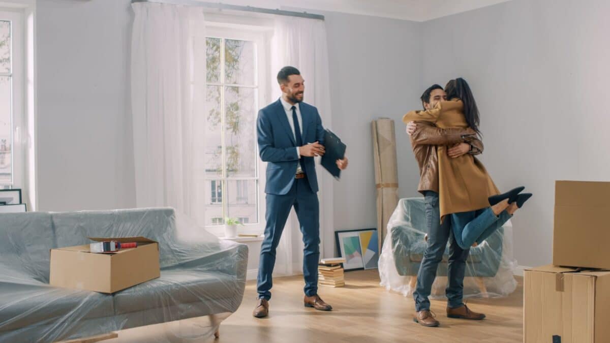 Woman jumping into a man's arms, celebrating while their real estate agent looks on.