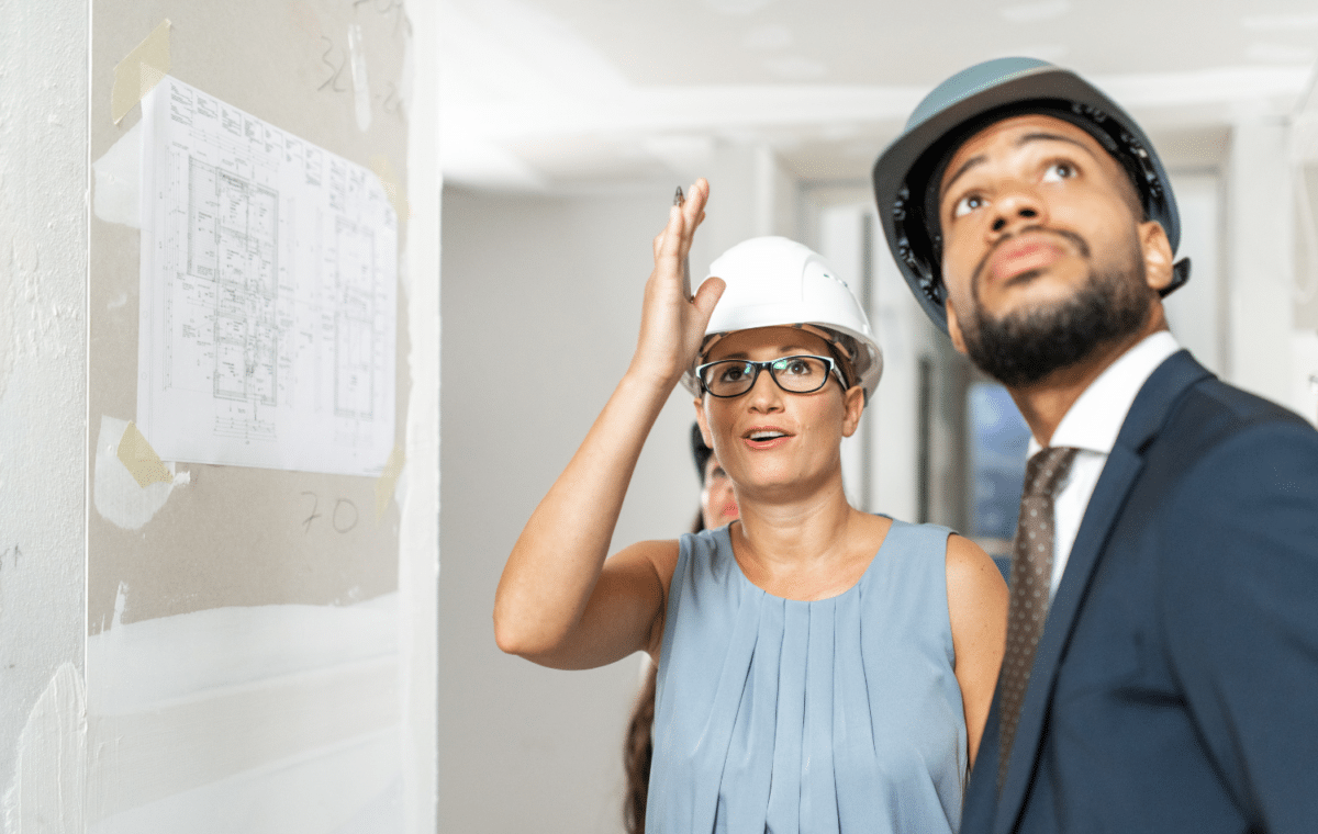 Woman with a hardhat looking at a blueprint and talking to a man with a hardhat.
